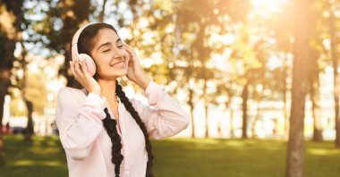 Kampüste dizüstü bilgisayarla dinlenen, kulaklık takan ve müzik dinleyen, teneffüste parkta bankta oturan mutlu Hintli öğrenci kız.
