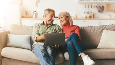 Cheerful Senior Couple Using Laptop Computer Sitting On Couch And Browsing Internet Together At Home. Mature People Use Gadget Watching Movie Online On Weekend. Full Length Shot