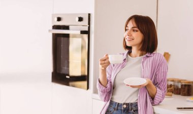 Günaydın ve lezzetli kahve, dinlen ve hafta sonunun tadını çıkar. Kafkasyalı genç bayan modern mutfakta kahve içiyor ve hayal kuruyor, boş alana bakıyor.