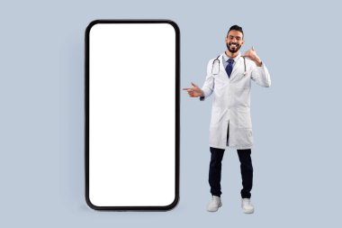 Arap erkek doktor büyük cep telefonu ekranını işaret ediyor ve kameraya gülümserken beni işaret ediyor. Mavi stüdyonun arka planında duruyor. Kare Çekim, Tıbbi Reklam İçin Model
