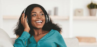 Positive attractive millennial african american woman in casual with wireless headset and cellphone sitting on couch at home, listening to music, looking at copy space and smiling