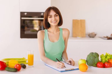Spor giyim kuşamında sporcu kadın günlük diyet ya da sağlıklı yemek tarifi yazıyor, mutfakta taze malzemelerle duruyor. Aktif sağlıklı yaşam tarzı, sağlıklı beslenme konsepti