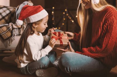 Mutlu noeller. Heyecanlı anne ve küçük kızı paketlenmiş hediye kutusunu açıyor dekore edilmiş oturma odasında sıcak zeminde oturuyor, tatilini evde kutluyor, arka planda bulanık ışıklar yanıyor.