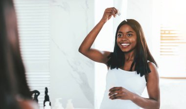 Cilt Kozmetik. Banyoda aynanın yanında dikilirken Düşürücü ile yüz serumu süren mutlu siyahi genç kadın, yansımasına gülümseyen, seçici odaklı