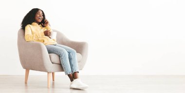 Evdeki koltukta oturan kahve fincanı ile sıradan giyinmiş mutlu genç siyahi bayan, sevgilisiyle veya arkadaşıyla telefon görüşmesi yapıyor, fotokopi odasına bakıyor ve gülüyor, panorama, beyaz arka plan