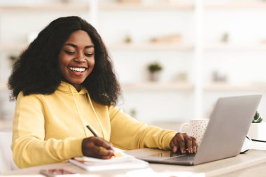 Evinden online derslere katılan zeki, çekici genç siyahi kadın çalışma masasında oturuyor, dizüstü bilgisayar kullanıyor ve notlar alıyor, kopyalama alanı yaratıyor. E-eğitim, eğitim, çevrimiçi kurs kavramı