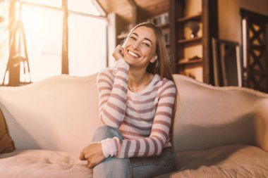 Neşeli, çekici genç bayan evde yalnız poz veriyor, günlük kıyafetler giyiyor, kanepede oturuyor ve gülümsüyor, kameraya bakıyor, rahat oturma odasına bakıyor, yakın çekim portresi, bedava fotokopi alanı