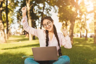 Bilgisayarında dizüstü bilgisayarla aşırı neşeli Hintli kız öğrenci dışarıda oturuyor, üniversite kampüsünde oturuyor, sınavı geçtiği için mutlu, başarıyı kutluyor.