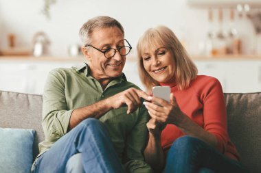 Yaşlı eşler cep telefonu kullanıyor, cep telefonu mesajlaşmasını işaret ediyor ve evdeki koltukta oturup interneti inceliyor. Sosyal medyada Akıllı Telefon Uygulamasıyla Olgun Eşler Ağı