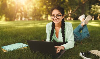 Açık havada okuyan mutlu Hintli kız öğrenci, çimenlikte dizüstü bilgisayarla uzanmak, ödev yapmak veya derslere hazırlanmak, kameraya gülümsemek, boş alan.
