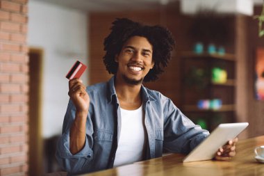 Neşeli gülümseyen kıvırcık milenyum siyahi adamın kredi kartı ve tableti kafenin içindeki ahşap masada online alışveriş yapıyor. Büyük satış, finans, harika haberler, uzaktan kumanda, duygu ve alımlar için uygulama