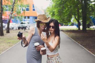 Dışarıda iki kız arkadaş akıllı telefonla selfie çekiyor. Boho şık moda kıyafetleri içinde genç bayan turistler, yaz parkında inliyor ve eğleniyorlar. Birlikte seyahat, yaşam tarzı portresi.