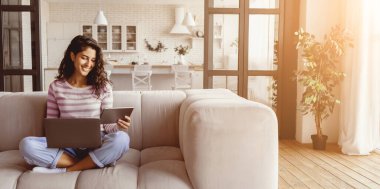 Genç bir kadın parlak bir oturma odasındaki kanepeye dizüstü bilgisayar ve tablet kullanarak rahatça oturur. Doğal ışık boşluğu doldurur, dinlenmek için sıcak ve davetkar bir atmosfer yaratır..