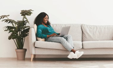 Tarz sahibi Afro-Amerikan milenyum bayan bağımsız müteahhit evden çalışıyor, rahat oturma odasındaki boş beyaz duvarın yanında oturuyor, modern dizüstü bilgisayar kullanıyor, erkeklerle sohbet ediyor, boşluğu kopyalıyor.