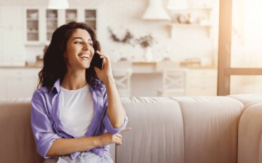 Genç bir kadın oturma odasındaki hafif bir kanepede oturuyor. O, telefonda neşeyle konuşuyor, mutluluk saçıyor. Sıcak güneş ışığı boşluğu doldurur, neşeli atmosferi güçlendirir..