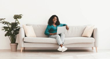 Neşeli Afro-Amerikan milenyum bayan bağımsız müteahhit evden çalışıyor, rahat oturma odasındaki boş beyaz duvarın yanında oturuyor, modern dizüstü bilgisayar kullanıyor, erkeklerle sohbet ediyor, boşluğu kopyalıyor.