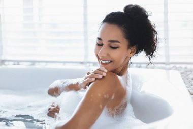 Köpük banyosunda sırılsıklam olan bir kadın. Penceresinden gelen parlak ışıkla modern bir banyoda. O dinlenirken su yavaşça etrafa sıçrıyor..