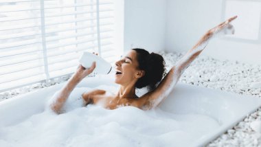 Bir kadın köpük banyosunda eğlenir, tek eliyle bir içki tutarken sevinçle kolunu kaldırır. Banyo doğal ışık ve beyaz dekorla dolu. Bu da sakin bir atmosfer yaratıyor..