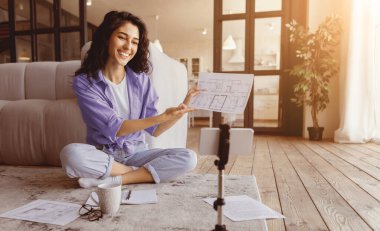 Genç bir kadın, video görüşmesi sırasında mimari planlarını gösterirken sırıtarak yerde bağdaş kurarak oturuyor. Kâğıtlar ve bir kahve fincanı yakınlarda, rahat ve parlak bir yere saçılmış..