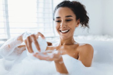 Bir kadın parlak bir gülümsemeyle köpük banyosundan hoşlanır. Ortam sakin, hafif ışıklandırma ve bol köpükle dolu. Bir banyo balosu düzenler, kendini şımartır..