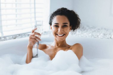 Köpük dolu bir köpük banyosunda oturan bir kadın gülümsüyor. Elinde bir bardak içki tutuyor, beyaz fayanslı ve şık bir atmosferi olan parlak bir banyoda bir dakikalık dinlenmenin tadını çıkarıyor..