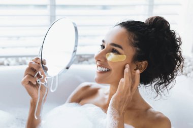 Bir kadın banyoda huzurlu bir anın tadını çıkarır, yansımasına gülümserken altın bir göz bandı takar. Etrafını saran baloncuklar, kendine bakmanın rahatlatıcı atmosferini artırıyor..