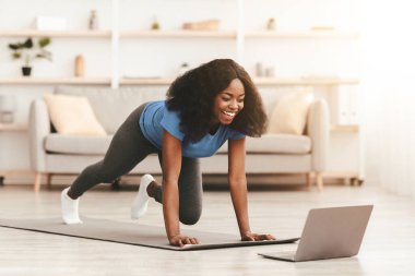 Pozitif genç Afro-Amerikan atletik kadın evde egzersiz yapıyor, dizüstü bilgisayar kullanıyor, spor salonunda spor salonunda spor yapan siyahi bayan spor yapıyor, vücut gücü üzerinde çalışıyor, yer kopyalıyor.