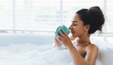 Bir kadın, elinde mavi bir fincan çayla baloncuk dolu bir banyoda huzur içinde oturuyor. Yumuşak güneş ışığı panjurların arasından süzülür, dinlenmek için mükemmel sakin bir atmosfer yaratır..