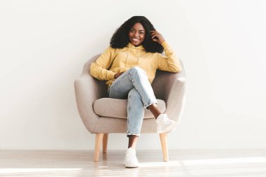 Çekici, gür saçlı, rahat giyimli, koltukta oturan, kameraya bakıp gülümseyen siyah bir kadın. Afro-Amerikan bir bayanın hafta sonunu yalnız geçirmesi çok güzel.