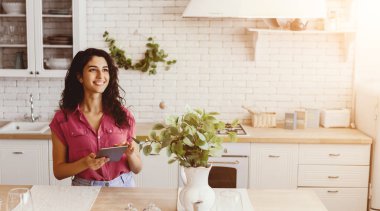 Güneşli bir mutfakta elinde tabletle gülümseyen bir kadın. Taze bitkiler mekanı süsler ve doğal ışık içeri akar, neşeli atmosferi güçlendirir..