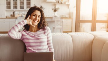 Kıvırcık saçlı genç bir kadın, rahat bir oturma odasında açık renkli bir kanepede rahatça oturur. Gülümsüyor ve sanki laptopuyla uğraşırken telefonla konuşuyormuş gibi hareket ediyor..