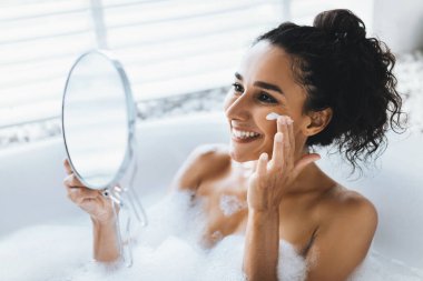 Genç bir kadın köpük dolu bir küvette oturuyor, yüzüne krema sürerken gülümsüyor. Küçük bir ayna tutuyor ve iyi aydınlatılmış bir banyo ortamında mutlu görünüyor..