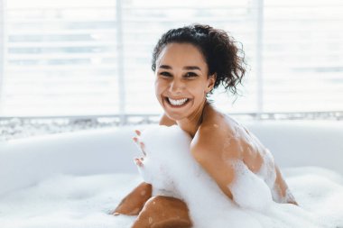 Bir insan köpük banyosundan zevk alır, köpükle çevrili iken neşeyle güler. Doğal ışık yakındaki pencerelerden süzülerek rahatlama ve kendine bakım için sakin bir ortam yaratıyor..