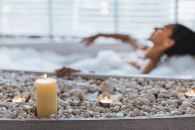 Bir kadın, mumlar ve banyo taşlarıyla çevrili huzurlu bir banyodan hoşlanır. Yumuşak ışık ve köpük rahatlatıcı bir ortam yaratır..