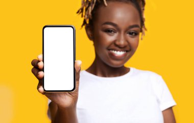Mobil uygulama kavramı. Sarı arka planda boş ekranlı akıllı telefon gösteren neşeli bir kadın. Beyaz ekran ile gülümseyen ve cep telefonu tutan Afrikalı Amerikalı genç bayana yakın çekim