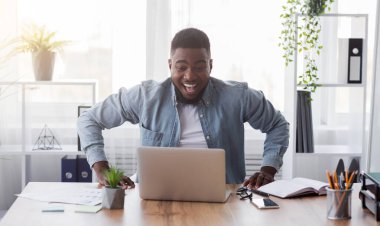 Maaş artışı. Neşeli Afro-Amerikan işçisi dizüstü bilgisayara heyecanla bakıyor. İnternetten iyi haberler aldıktan sonra mutlu oluyor. Maaşına zam yapılıyor..