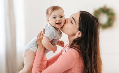 Anneler sever. Mutlu, genç bir anne, çocuğuyla evde oynayıp eğlenirken elini tutup yanaktan öpüyor. Doğum günün kutlu olsun.