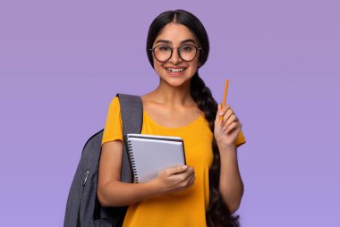 Cevabı biliyorum. Güzel gülümseyen gözlüklü Hintli bayan öğrencinin portresi. Elinde not defteri, elinde kalemle mor stüdyo arka planına izole edilmiş sırt çantası.