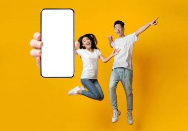 Harika bir teklif. Heyecanlı Asyalı çift, ellerinde boş beyaz hücre ekranı, neşeli adam ve atlayan bayan, sarı portakal rengi stüdyo arka planı, sahte kolaj pankartı olan büyük akıllı telefonu tutuyor.