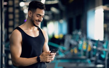 Spor Kulübü 'nde eğitim görürken akıllı telefon kullanan yakışıklı, gülümseyen Arap Atletizm Mesajlaşması ya da spor salonunda zaman geçirirken yeni spor uygulaması taraması yapan, uzay kopyalayan genç bir orta doğulu.