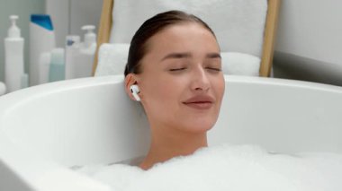 Bir kadın köpük banyosunda sırılsıklam, gözleri kapalı, sakin ve rahat görünüyor. O, yumuşak beyaz köpükle çevriliyken kablosuz kulaklık takıyor. Banyonun rahatlatıcı bir atmosferi var..