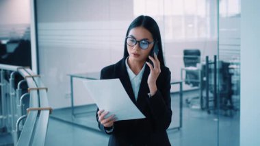 Çağdaş bir kurumsal çalışma alanında, iş kıyafeti giymiş Asyalı bir kadın telefonuyla konuşurken önemli belgeleri gözden geçirir, yoğun bir ortamda mesleki davranışını gösterir..