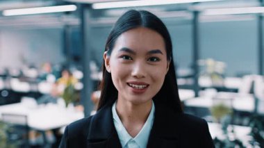 Profesyonel bir Asyalı kadın, modern bir şirket ortamında çalışırken kendine güvenerek gülümser. Meşgul ve iş yerinde ekip üyeleriyle işbirliği yapmaya hazır görünüyor..