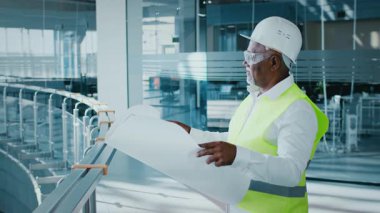 A mature African American businessman, dressed in a white shirt and safety gear, reviews architectural plans in a modern corporate office space during the day.