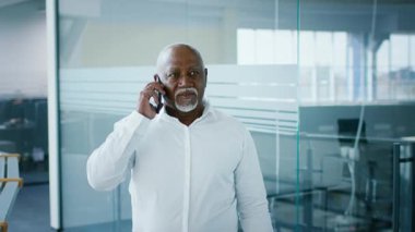 A mature African American man in a white shirt is on the phone in a bright, contemporary office. He appears focused and confident while discussing business matters on his smartphone.