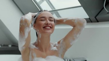 Bir kadın saçını köpük banyosunda yıkarken neşeyle gülümser. Modern banyo tavan pencerelerinden gelen doğal ışıkla aydınlanıyor ve huzurlu bir atmosfer yaratıyor..