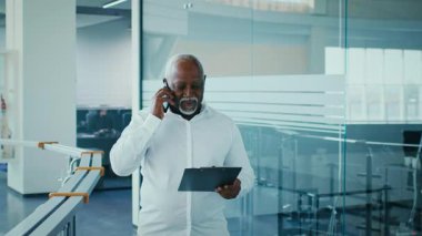 A mature African American man in a white shirt talks on the phone while holding a clipboard in a contemporary office environment. The setting features glass walls and a professional atmosphere.