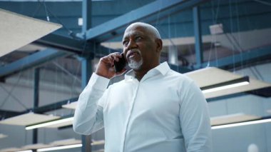 A mature African American man in a white shirt speaks on his phone while standing in a stylish office. He appears focused and professional, embodying a corporate lifestyle.