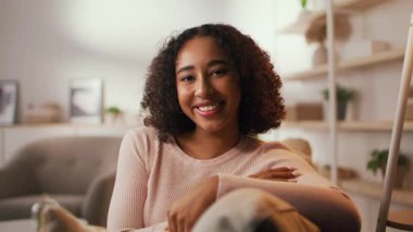 Kıvırcık saçlı genç bir Afrikalı Amerikalı kadın evinde rahat bir şekilde oturuyor, sıcak bir şekilde gülümsüyor. Konforlu ortam, rahat bir yaşam tarzını vurgulayarak yumuşak ışıklandırma ve şık bir dekor içeriyor..