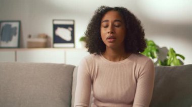 Genç bir Afrikalı Amerikalı kadın kanepede rahatça oturup hapşırıyor ve hastalanıyor. Konforlu yaşam alanı yavaşça aydınlatılıyor. Rahatlamış bir atmosfer yaratıyor..